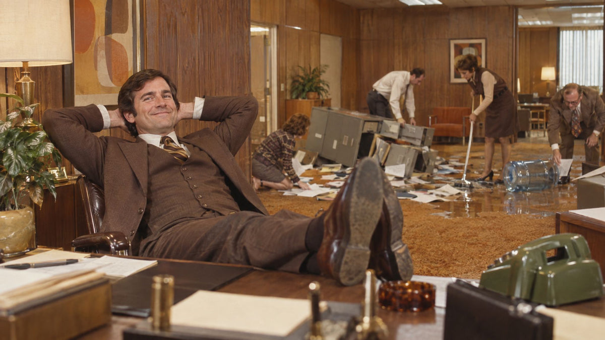 Smiling man in a brown suit with his feet on the desk while colleagues clean up an office mess behind him