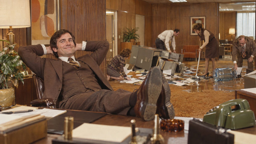 Smiling man in a brown suit with his feet on the desk while colleagues clean up an office mess behind him
